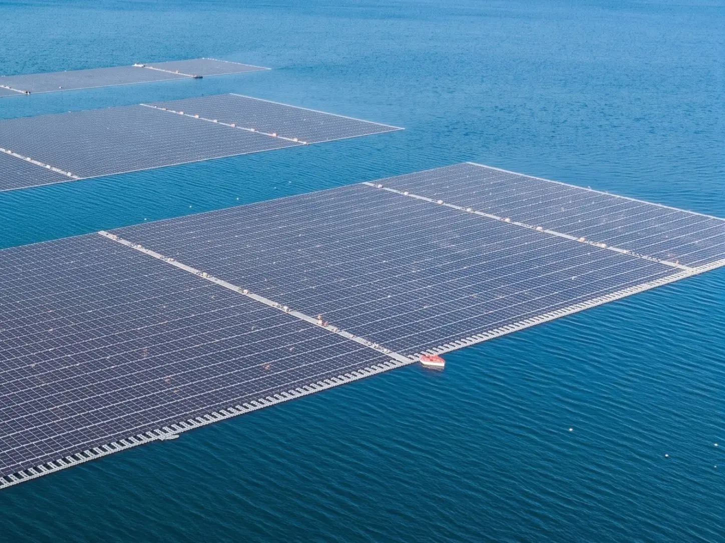 Aerial view of large floating solar arrays on open blue water with central walkways and a service boat at the array edge