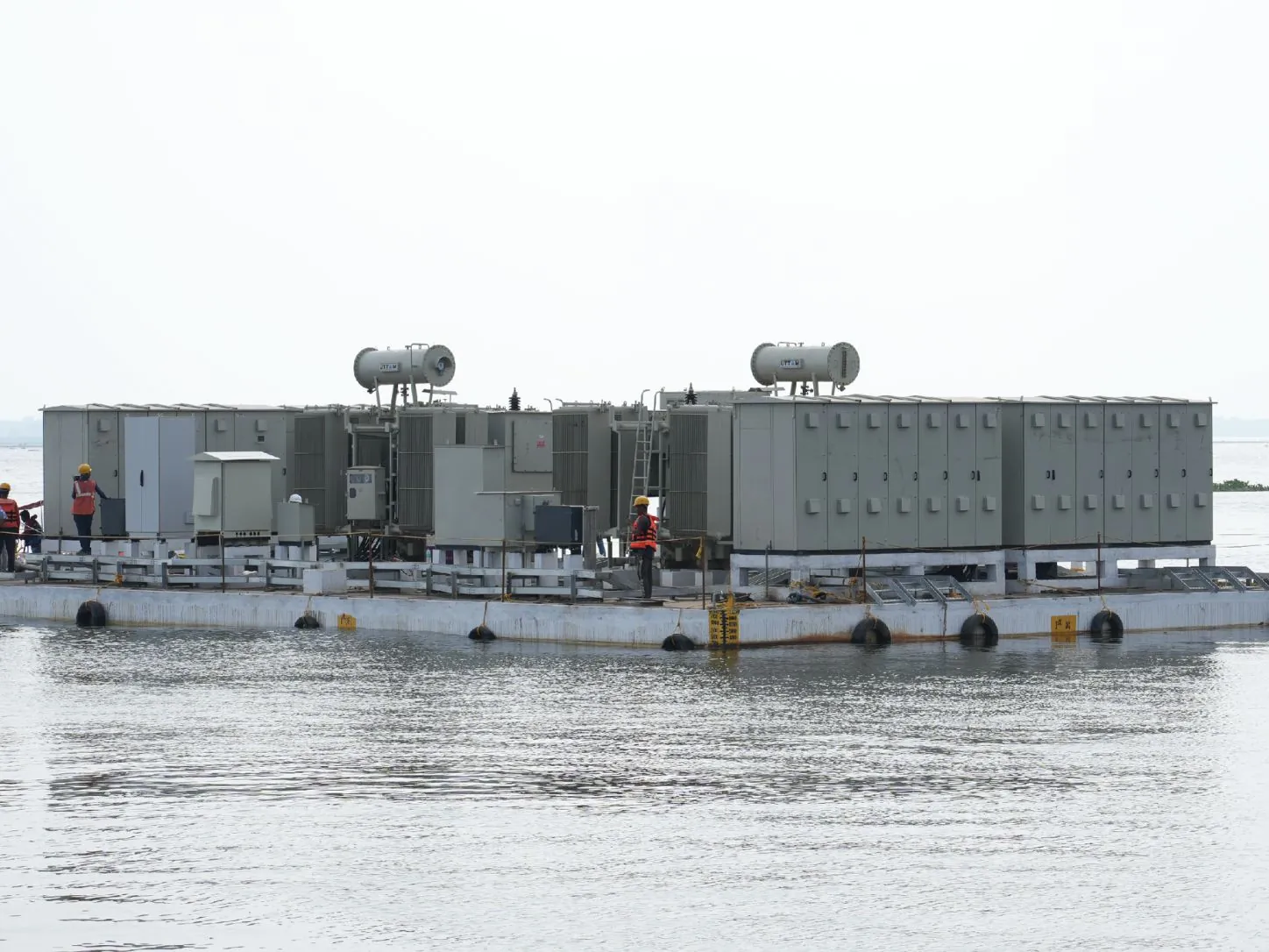 Floating electrical platform carrying transformers and switchgear cabinets on a reservoir, with technicians in life jackets on deck